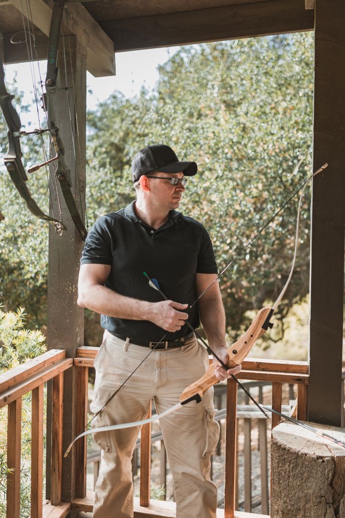 Concentrated young male in casual clothes and cap aiming while shooting with bow and arrow in archery club in forest